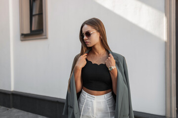 Wall Mural - Elegant beautiful young business woman with vintage sunglasses in fashionable urban clothes with a green blazer stands near a white wall on the street