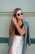 © alones - Fashionable beautiful young woman model with vintage sunglasses in stylish clothes with a blazer stands near a green wall and straightens her hair on the street