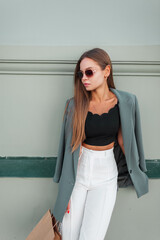 Wall Mural - Elegant beautiful young woman model with sunglasses in fashion clothes with blazer sitting near a green wall in the city. Urban chic lady