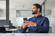 © insta_photos - Smiling professional Indian businessman employee looking aside holding smartphone sitting at desk. Happy busy business man executive using cell phone thinking of new mobile solutions working in office