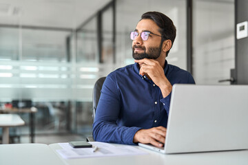 Happy busy Indian businessman worker thinking of future ai solutions looking aside working on laptop. Smiling professional business man using computer planning financial strategy at work in office.