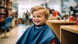 © ND STOCK - A 3-year-old boy wearing a protective cape sits in a happy barber's chair while getting a haircut in a cutely decorated shop for children.