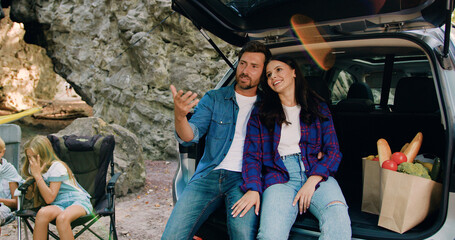 Loving couple sitting on car trunk and child resting in shade of tree near the car in nature have picnic. Happy smiling family man woman and kids traveler vacation countryside. Road trip together