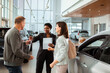 © Marko Geber - Couple signing paperwork to purchase a new car at dealership with saleswoman