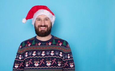 Wall Mural - Handsome bearded caucasian guy in sweater and Santa hats stands on blue background in studio and teeth smiling. Portrait european guy with Christmas mood. Holiday banner. New Year.