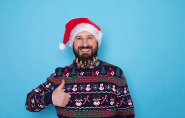 Positive bearded man in holiday sweater and Santa hat showing thumb up, smiling and looking at camera on blue studio background. Millennial man promoting Christmas sale or special offer. Xmas.