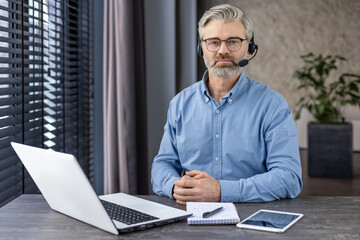 Wall Mural - Portrait of serious confident businessman with headset phone for video call, mature gray haired man looking concentrated at camera, boss at workplace inside office with laptop.