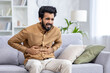 © Liubomir - A young Indian man is sitting on the couch at home and holding his stomach with his hands, feeling severe spasm and discomfort. Winced in pain.