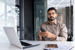 © Liubomir - Portrait of successful Indian businessman, serious thinking man looking at camera at workplace inside office, boss holding phone, using laptop.