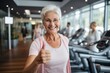 Portrait of smiling senior woman on exercise bike in fitness center with people on background