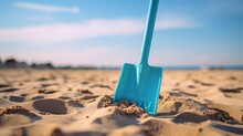 A Plastic Toy Shovel With A Handle Is Stuck In The Beach Sand. In The Background, You Can See The Sea. The Weather Is Sunny.