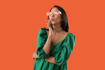 Poster - Beautiful stylish Asian woman in sunglasses blowing kiss on orange background