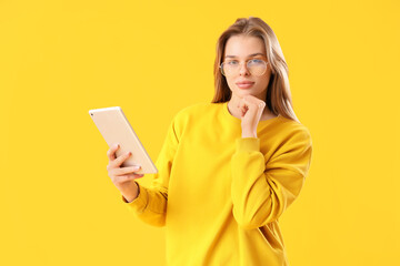 Wall Mural - Thoughtful young woman in eyeglasses using tablet computer on yellow background