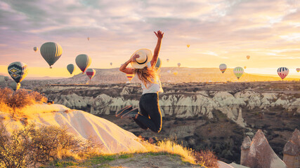 Wall Mural - Young female tourist jumping in the air with hot air balloons background at sunrise in Cappadocia, Turkey
