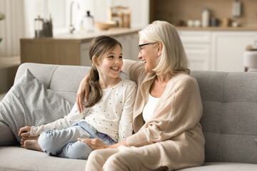 Wall Mural - Cheerful loving grandmother and cute grandkid girl talking and laughing on sofa, enjoying funny conversation, trust, family leisure. Grandma speaking to child girl at home