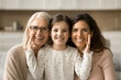 © fizkes - Cheerful cute little girl, mom, grandmother sitting close indoors, hugging, looking at camera with toothy smiles. Happy daughter kid touching cheeks of younger mother and grandma head shot portrait