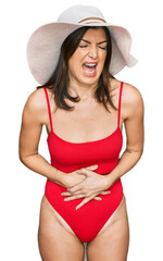 Beautiful brunette woman wearing swimsuit and summer hat with hand on stomach because nausea, painful disease feeling unwell. ache concept.