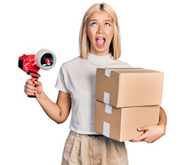 Sticker - Beautiful young blonde woman holding packing tape and cardboard boxes angry and mad screaming frustrated and furious, shouting with anger looking up.