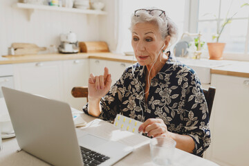 Telemedicine concept. Senior female doctor consulting her patients online, explaining routine of pills intake holding blister package in hands, wearing glasses on forehead using laptop and earphones