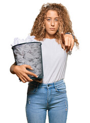 Sticker - Beautiful caucasian teenager girl holding paper bin full of crumpled papers pointing with finger to the camera and to you, confident gesture looking serious