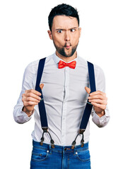 Poster - Hispanic man with beard wearing hipster look holding suspenders making fish face with mouth and squinting eyes, crazy and comical.