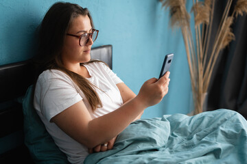 Beautiful young woman in glasses reading a message scrolling through social networks while sitting in bed in the morning at home