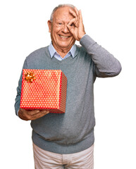 Canvas Print - Senior caucasian man holding gift smiling happy doing ok sign with hand on eye looking through fingers