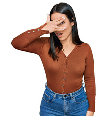 Wall Mural - Young hispanic woman wearing casual clothes peeking in shock covering face and eyes with hand, looking through fingers with embarrassed expression.