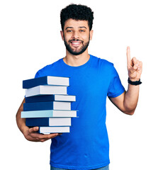 Sticker - Young arab man with beard holding a pile of books smiling with an idea or question pointing finger with happy face, number one