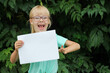 Blonde little girl with glasses holding a blank sheet of paper with space for text, the child against the background of green leaves.