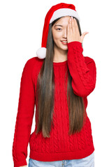 Poster - Young chinese woman wearing christmas hat covering one eye with hand, confident smile on face and surprise emotion.