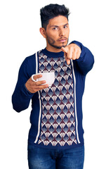 Wall Mural - Handsome latin american young man drinking a cup of coffee pointing with finger to the camera and to you, confident gesture looking serious