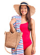 © Krakenimages.com - Young beautiful latin girl wearing swimwear and summer hat holding beach towel and bag looking positive and happy standing and smiling with a confident smile showing teeth