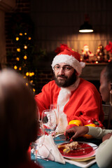 Wall Mural - Santa claus man in costume sitting at homr with diverse friends and family, enjoying festive dinner on christmas eve holiday. Young adult with hat spreading positivity during december holiday.