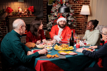 Wall Mural - Man in santa claus costume sitting at table with diverse people, enjoying festive dinner on christmas eve festivity. Young person in suit spreading positivity during december holidays.