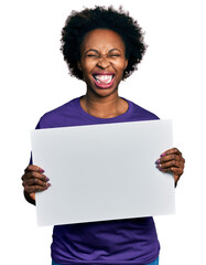 Canvas Print - African american woman with afro hair holding blank empty banner sticking tongue out happy with funny expression.