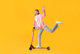 Portrait of excited businesswoman riding kick scooter and waving hand on yellow background