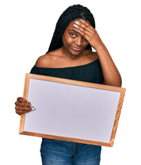 Poster - Young african american woman holding empty white chalkboard stressed and frustrated with hand on head, surprised and angry face