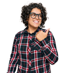 Canvas Print - Young hispanic woman with curly hair wearing casual clothes and glasses smiling with happy face looking and pointing to the side with thumb up.