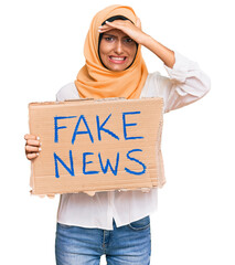 Poster - Young brunette arab woman holding fake news banner stressed and frustrated with hand on head, surprised and angry face