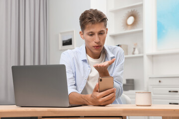 Poster - Happy young man having video chat via smartphone and blowing kiss at wooden table indoors. Long-distance relationship