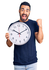 Poster - Young hispanic man holding big clock screaming proud, celebrating victory and success very excited with raised arms