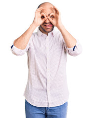 Poster - Young handsome man wearing elegant shirt doing ok gesture like binoculars sticking tongue out, eyes looking through fingers. crazy expression.