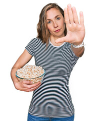 Sticker - Young blonde woman eating popcorn with open hand doing stop sign with serious and confident expression, defense gesture