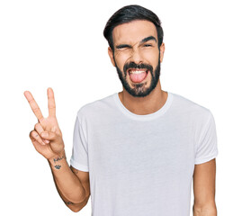 Wall Mural - Young hispanic man wearing casual white t shirt smiling with happy face winking at the camera doing victory sign. number two.