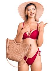 Sticker - Young beautiful woman wearing bikini and hat holding summer wicker handbag pointing thumb up to the side smiling happy with open mouth