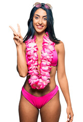 Wall Mural - Beautiful hispanic woman wearing bikini and hawaiian lei showing and pointing up with fingers number two while smiling confident and happy.