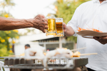 Outdoor barbecue with various meats on grill, two people holding plates and drinks, smoke rising, festive and casual atmosphere. holding glass of beer. family party.