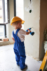 Wall Mural - Boy in work overalls drilling wall with toy electric drill tool at home during renovation. Kid in safety helmet using drilling instrument while working on apartment refurbishment.