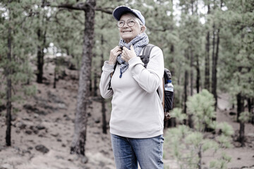 Wall Mural - Active senior woman holding backpack standing in the pines forest enjoying nature and adventure. Sport and healthy lifestyle concept